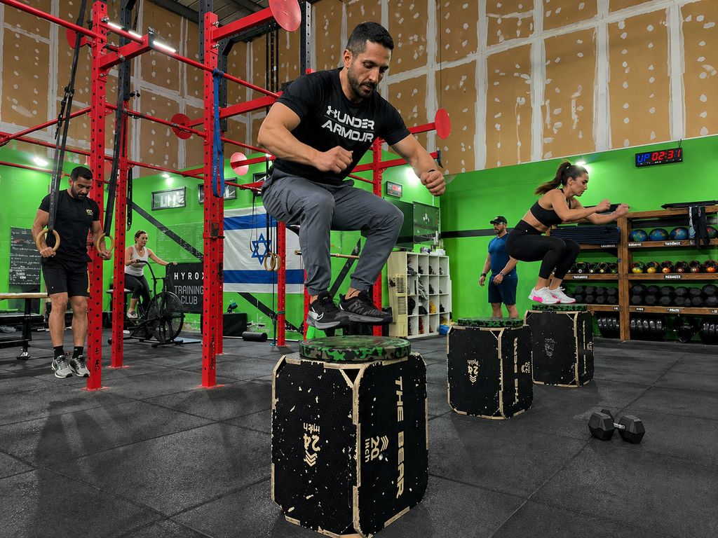 קבוצת מתאמנים מבצעת box jumps ותרגילי כוח באימון קרוספיט אינטנסיבי בתוך בוקס עם ריגים אדומים, קיר ירוק וציוד פונקציונלי ברקע.
